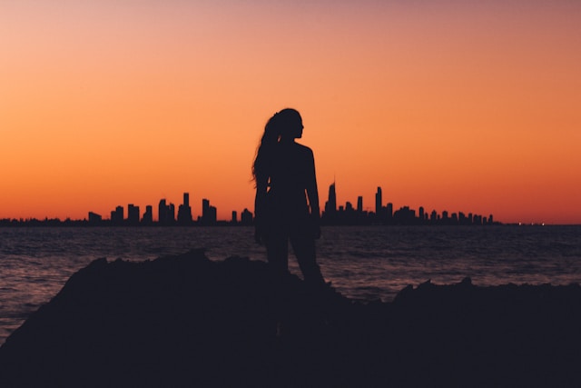 Silhouette of a woman standing by the ocean at sunset with the Gold Coast skyline in the background.